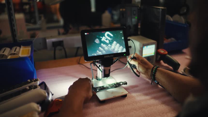 A technician meticulously examines a printed circuit board under a digital microscope in a dimly lit electronics workshop