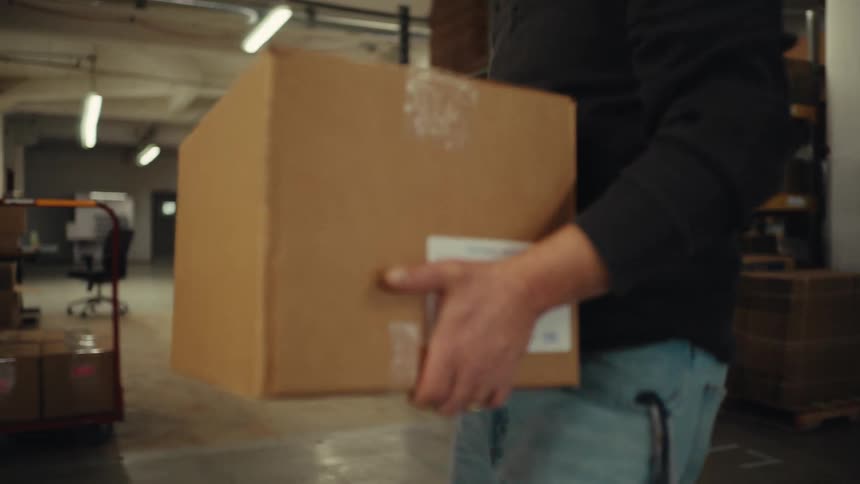 A warehouse worker picks a cardboard box off a shelf and carries it through the aisles in slow motion
