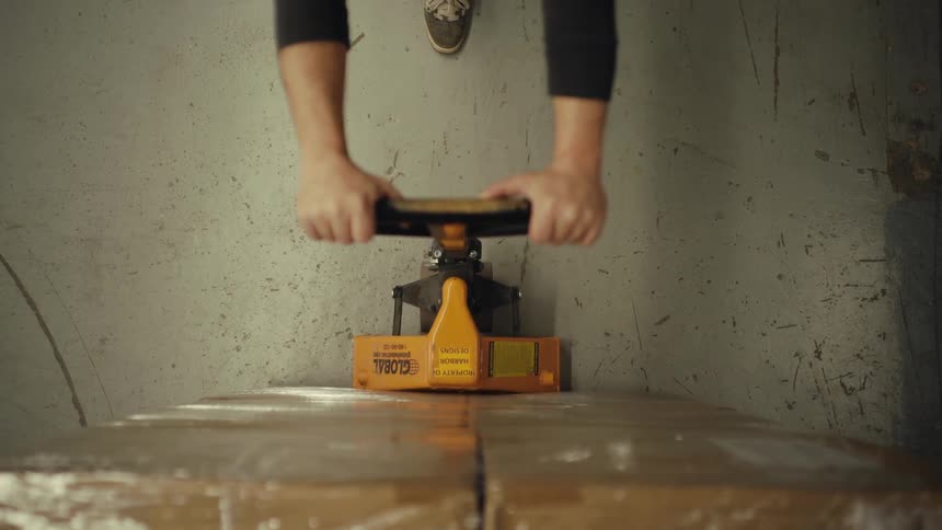 A top-down point of view from a pallet of boxes as a worker operates a pallet jack to lift and move the load in a warehouse