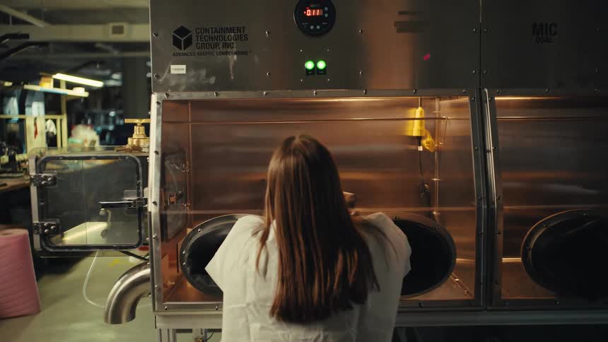 A female technician seen from behind operates a large stainless steel glovebox in a modern laboratory or manufacturing facility