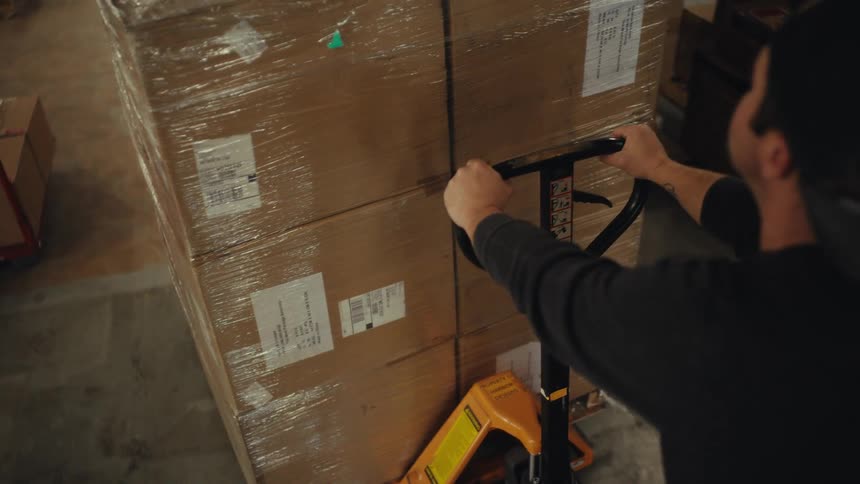 Over-the-shoulder shot of a warehouse worker pushing a pallet jack loaded with shrink-wrapped boxes