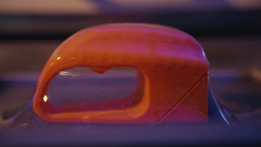 Macro shot of a 3D printer creating a vibrant orange object from a liquid polymer resin under ultraviolet light