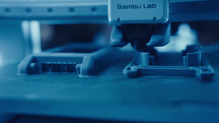 A close-up view shows a modern 3D printer meticulously fabricating a complex plastic part under cool blue lighting