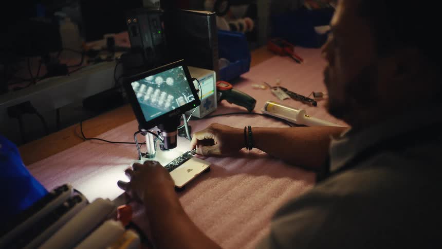 An electronics technician carefully inspects a circuit board under a digital microscope in a dimly lit workshop