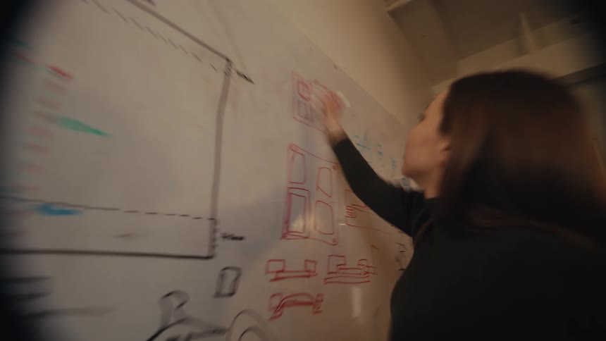 A focused female designer sketches technical diagrams and creative concepts on a whiteboard in a modern office
