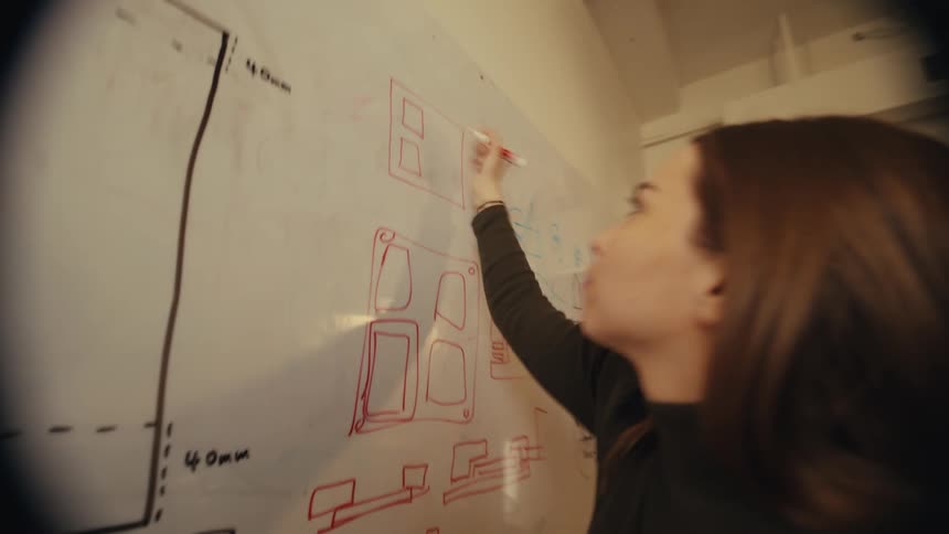 A focused female designer sketches out new product ideas with a red marker on a whiteboard in a modern office