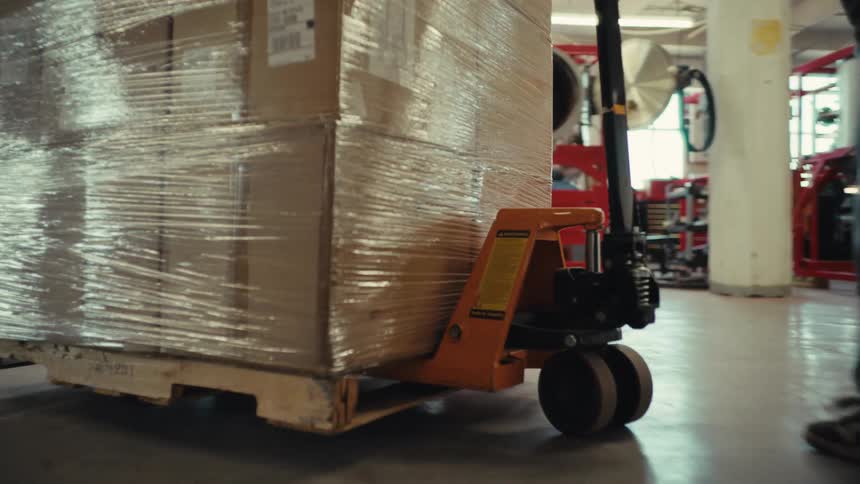 Low-angle slow motion of a worker pushing a pallet jack with shrink-wrapped cardboard boxes through a warehouse