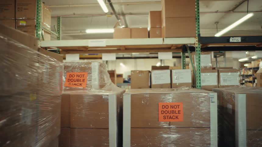 A slow pan reveals stacks of shrink-wrapped cardboard boxes with warning labels on industrial shelves in a large warehouse