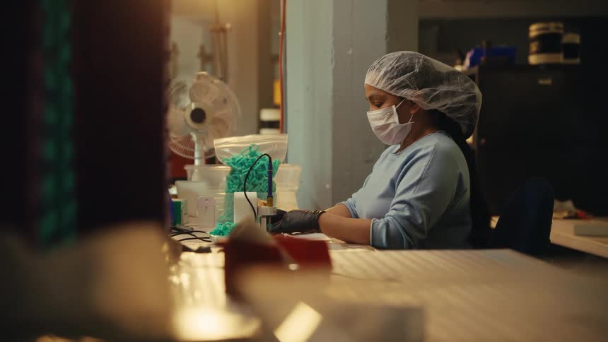 A female worker in protective gear carefully works on assembling a small product at a dimly lit factory workstation