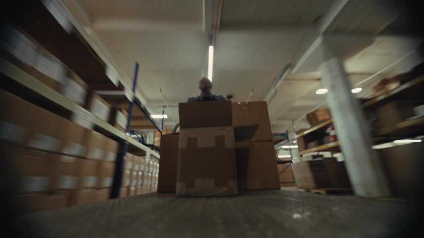POV shot from a dolly loaded with cardboard boxes being pushed through a busy warehouse by a male worker
