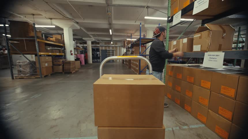 A male warehouse employee loads cardboard boxes onto a hand truck in slow motion, shot from the dolly's perspective