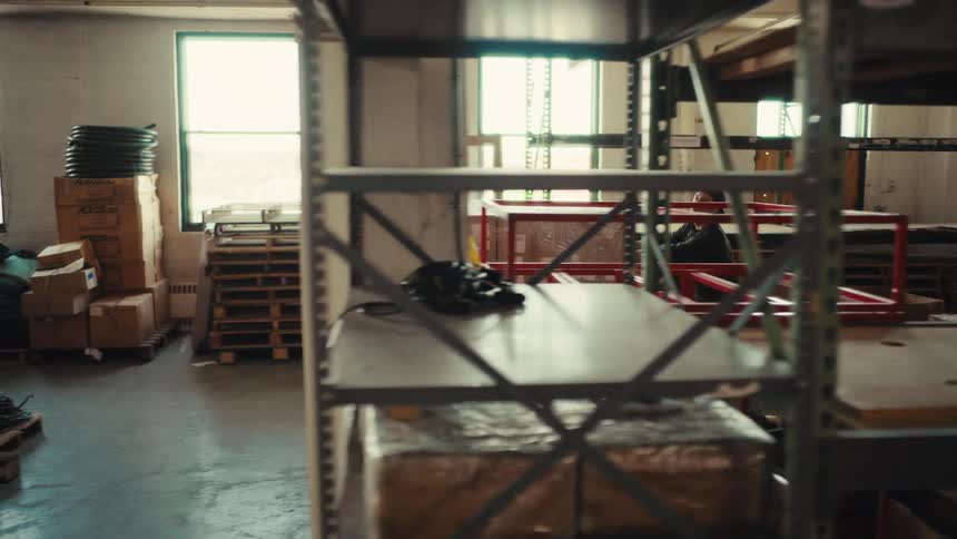 A warehouse worker moves a large pallet of boxes with a pallet jack through the aisles in a cinematic slow motion tracking shot