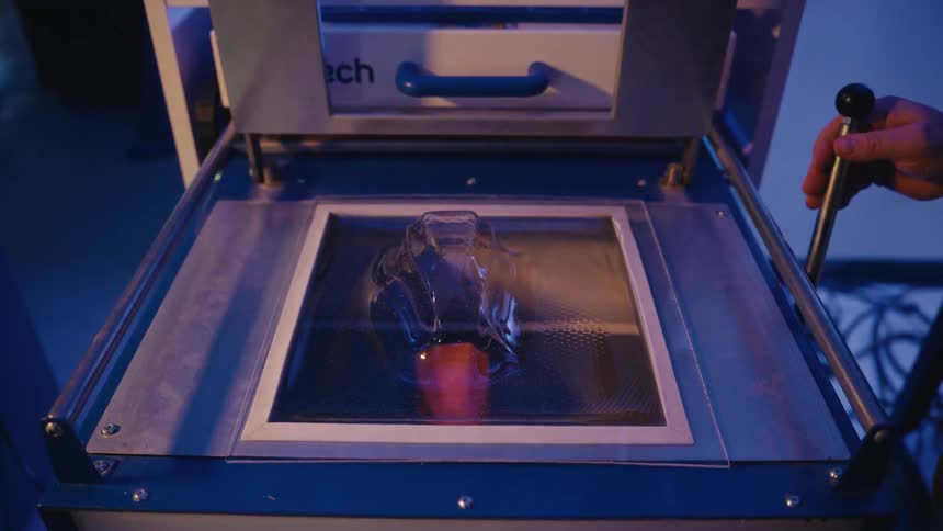 A thermoforming machine creates a clear plastic mold by vacuum sealing a heated sheet over an orange prototype in a workshop