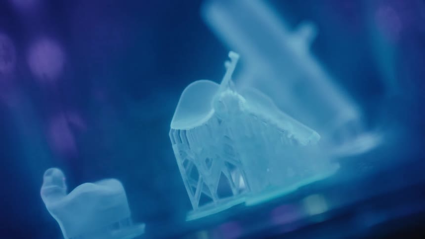 A close-up of a 3D printer creating a translucent dental implant model under a glowing blue light
