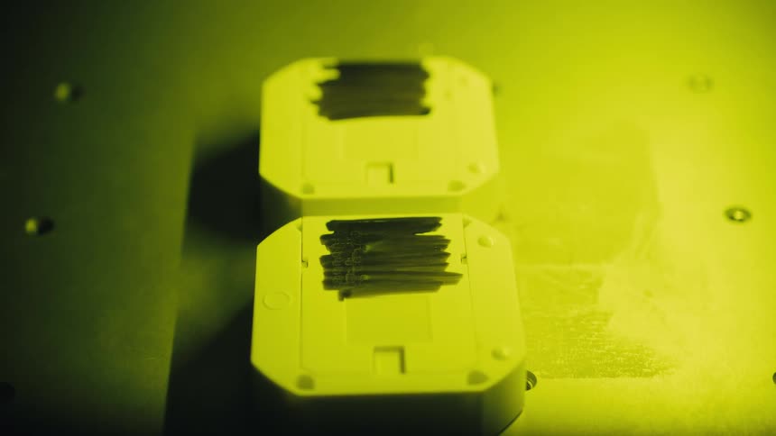 A precision laser engraver machine methodically marks numbers onto two white plastic components under a yellow safety light