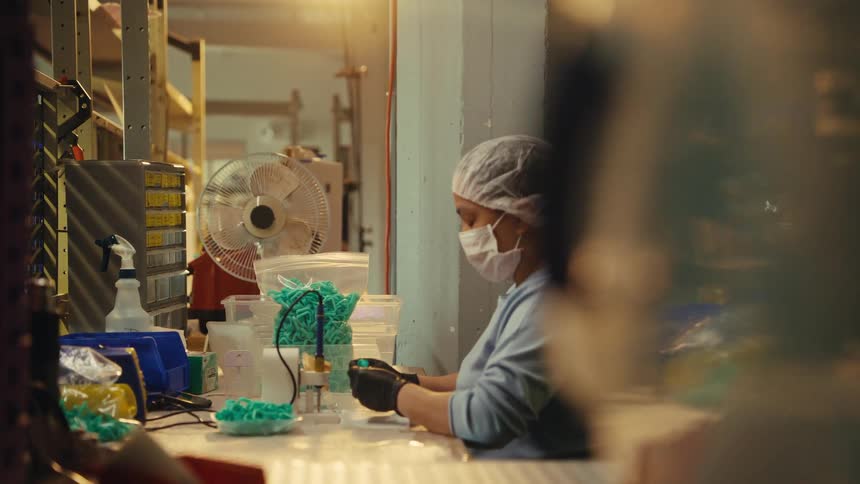 A female factory worker in a hairnet and mask carefully assembles small plastic parts at her workstation