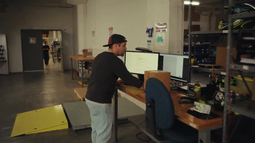 A male warehouse employee stands at his dual-monitor computer desk to weigh and process a shipping label for a package
