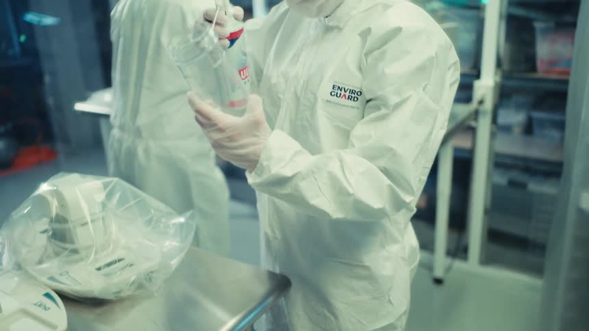 A lab technician in a full cleanroom suit operates machinery before meticulously cleaning a container with compressed air