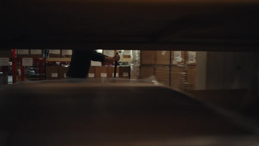 A warehouse worker moves a large pallet of shrink-wrapped boxes with a manual pallet jack through the aisles of a large industrial facility