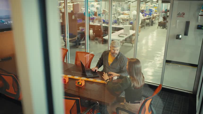 A man and woman collaborate on a new product design in a glass conference room overlooking a bustling manufacturing facility