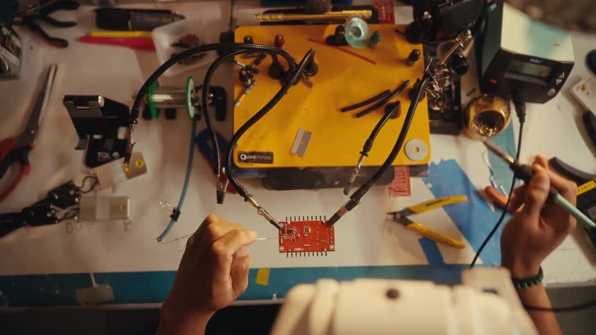 A top-down view of a technician's hands carefully soldering a red circuit board on a cluttered workbench