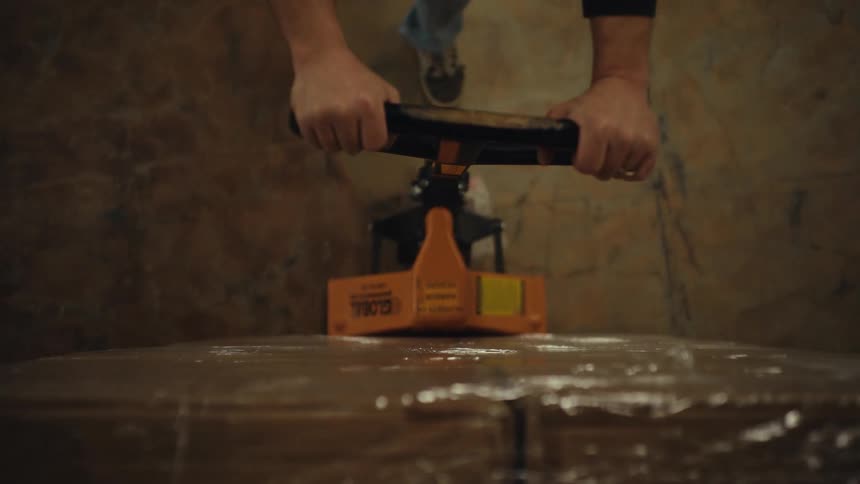 First-person point-of-view of a warehouse worker pulling a pallet jack stacked with shrink-wrapped boxes through a storage facility