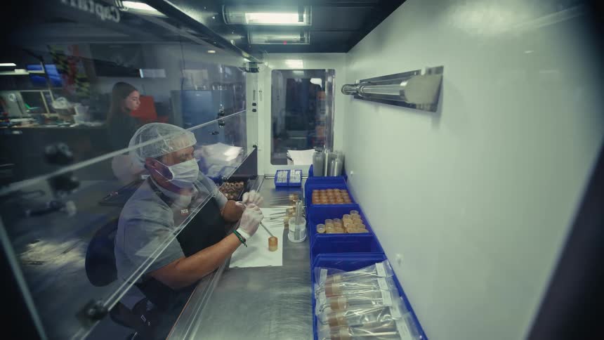 A worker wearing a face mask and hairnet carefully assembles small vials inside a sterile laboratory clean room hood