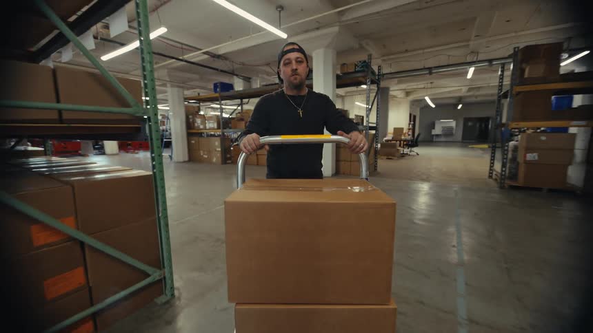 A male warehouse worker loads a cardboard box onto a dolly and pushes it down an aisle filled with shelves of inventory