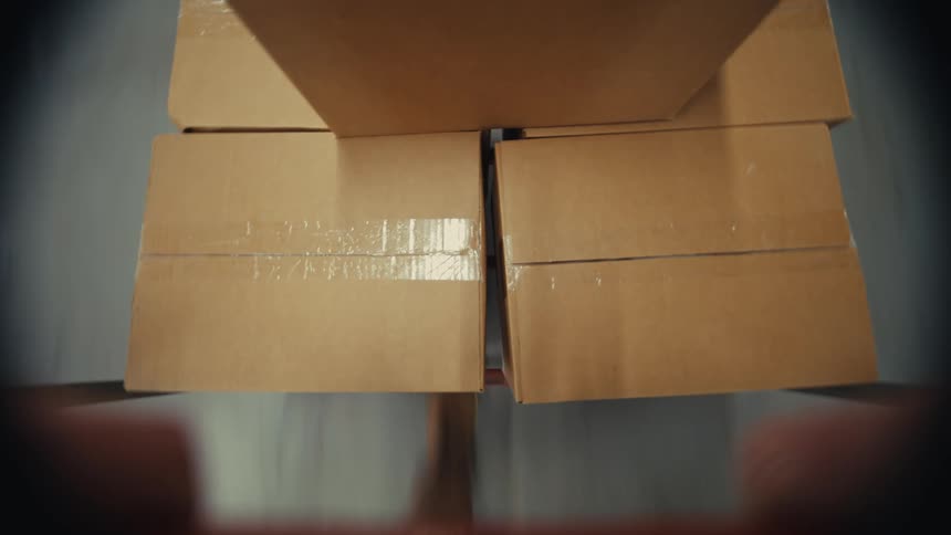 A first-person point of view of a warehouse worker pushing a hand truck loaded with sealed cardboard boxes