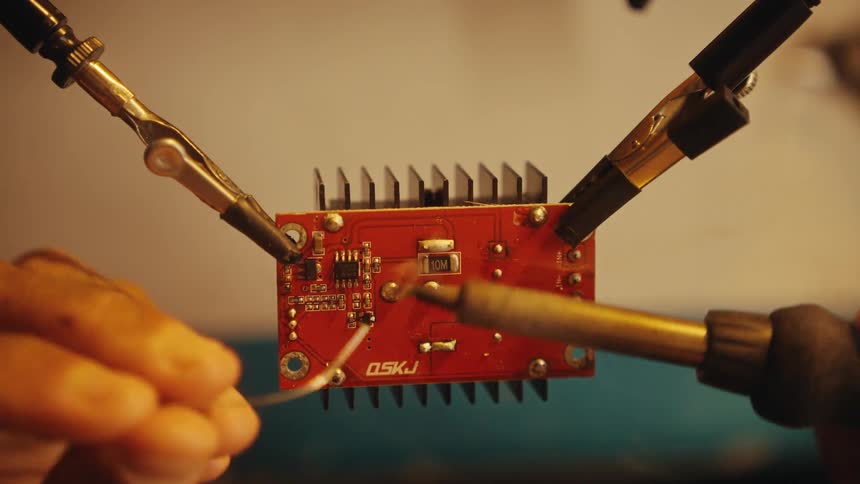 A technician carefully solders wires and components onto a red circuit board held in place by helping hands