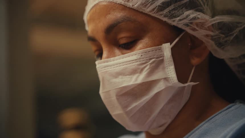 An extreme close-up profile shot of a female surgeon wearing a mask and hairnet, looking down with intense concentration