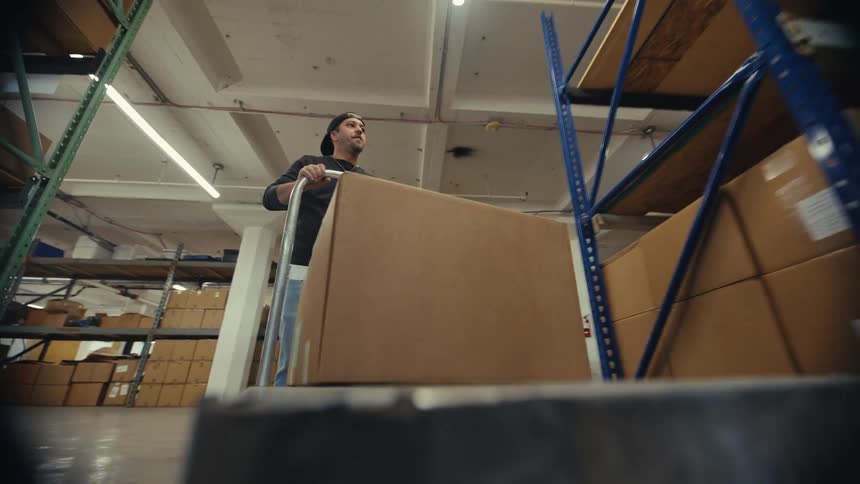 A warehouse worker loads a large cardboard box onto a dolly and pushes it down the aisle in a point-of-view shot