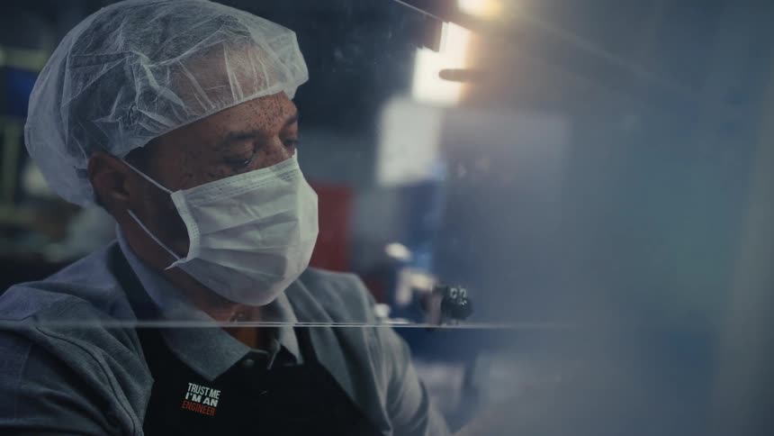 A masked engineer in a cleanroom meticulously assembles a complex mechanical component behind a protective screen