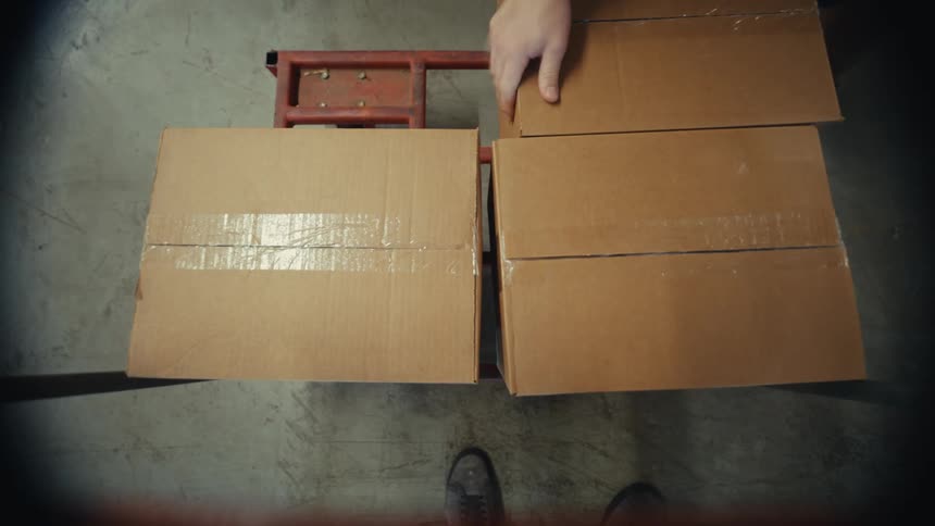 A first-person view of a worker loading cardboard boxes onto a red dolly and wheeling them across a concrete floor