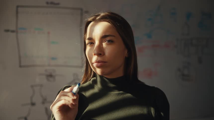 A determined young professional bathed in dramatic sunlight thinks through a complex problem on a whiteboard before turning to face the camera