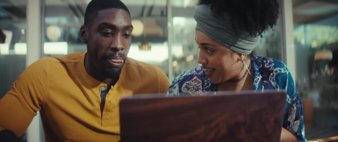 A young Black man and woman sit together at a table, intently looking at a laptop screen and discussing what they see