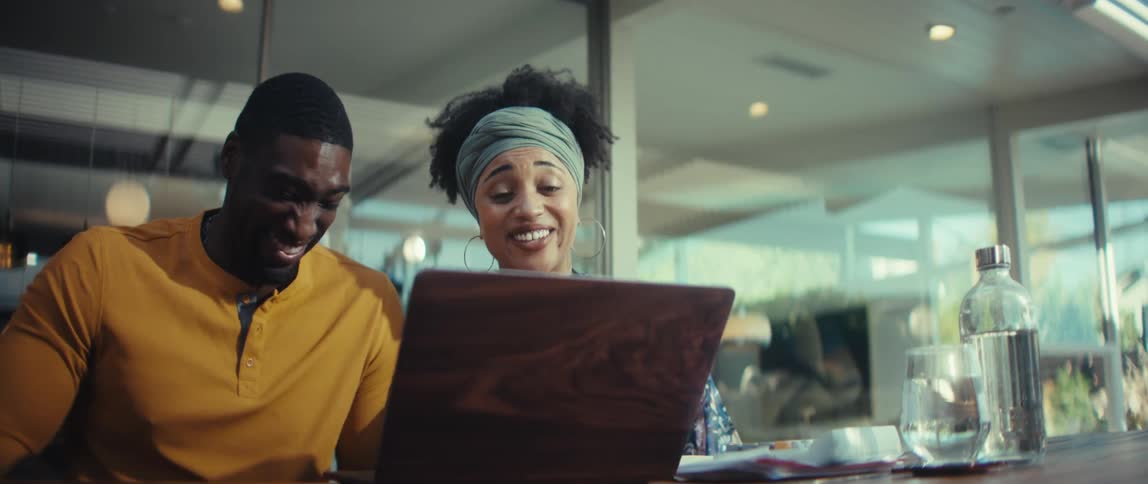 A joyful African American couple sits at a table, laughing and pointing at their laptop screen during a fun video call