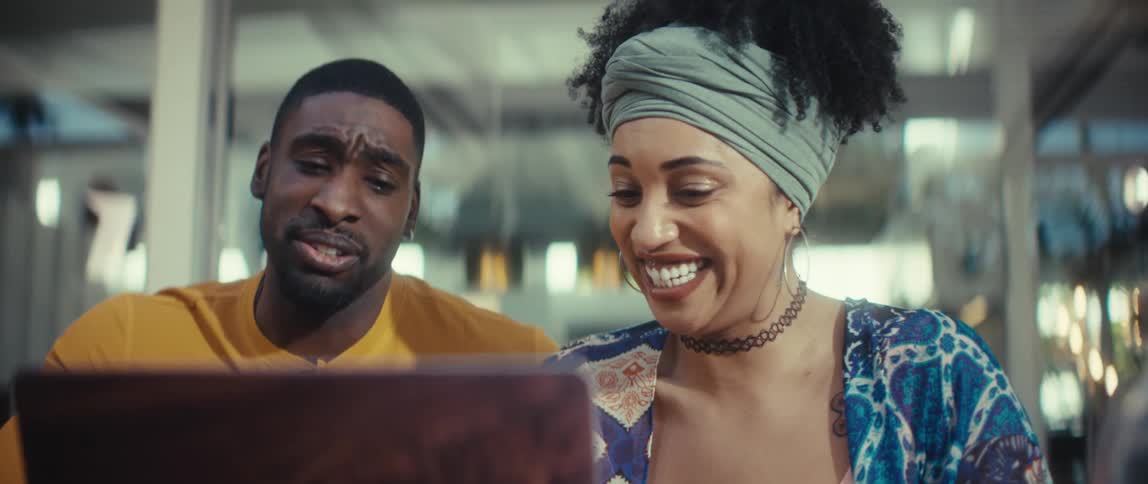 A stylish young man and woman have an enthusiastic discussion while looking at a laptop in a modern cafe