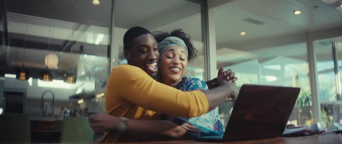 An ecstatic young couple celebrates getting good news on their laptop, cheering and hugging in slow motion