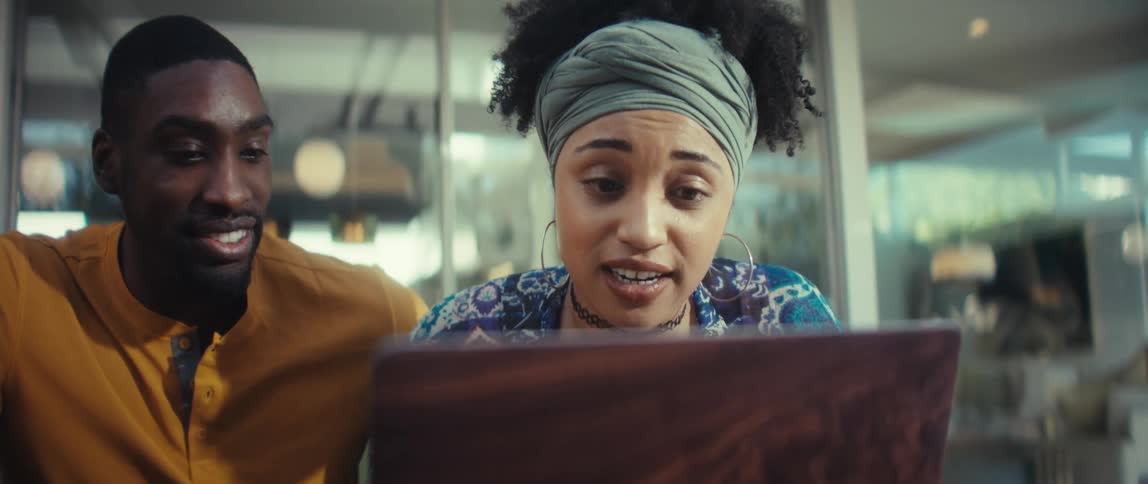A young, stylish Black couple gets excited while looking at a laptop together in a modern, sunlit cafe