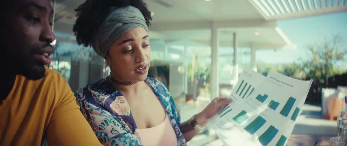 A young Black couple collaborates on their finances, with the man using a calculator and the woman explaining charts and graphs in their modern home