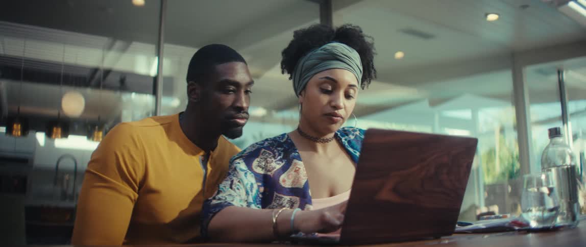 A young African American woman opens her laptop as her partner looks on, collaborating on a project from their modern home