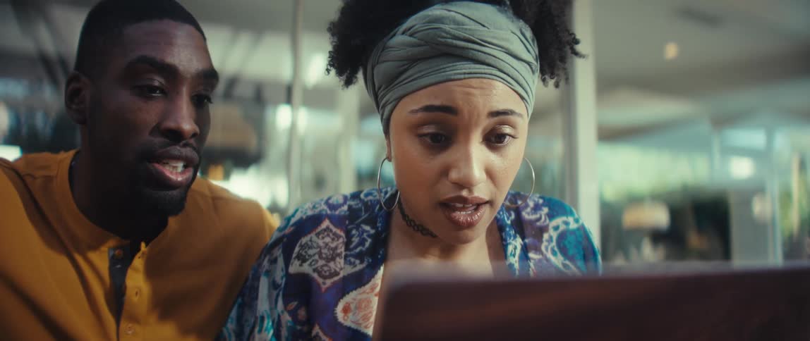A young Black man and woman are happily surprised while working together on a laptop in a modern cafe