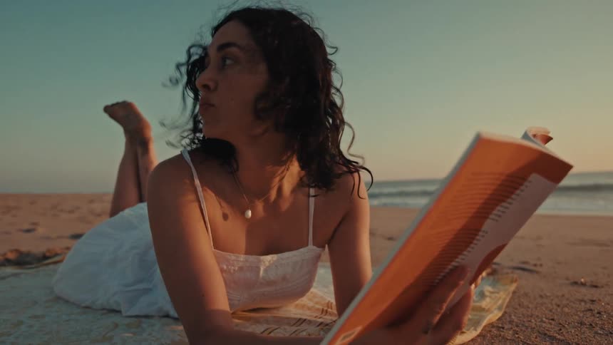 A beautiful young woman in a white dress relaxes on a blanket at the beach, peacefully reading a book at sunset