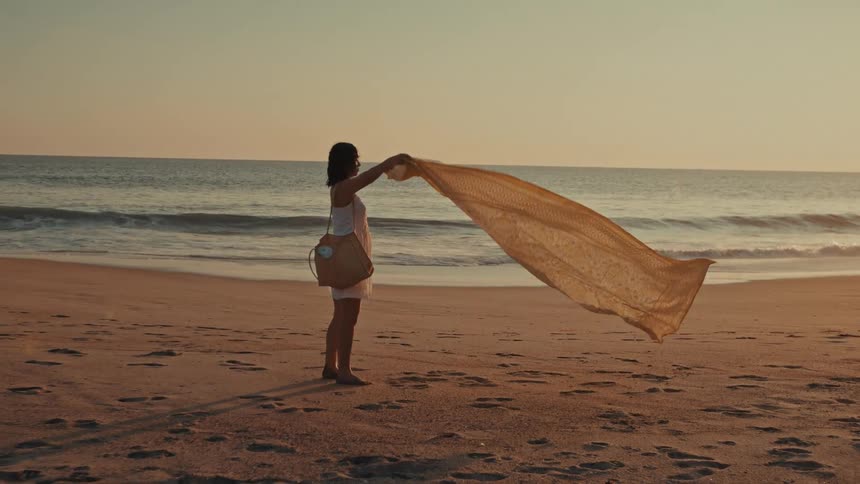 A woman standing on a sandy beach at sunrise holds up a scarf to catch the gentle ocean breeze in slow motion