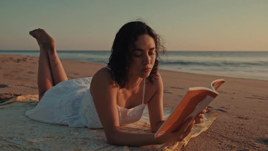 A young woman in a white dress reads a book while lying on a blanket on the beach at sunset