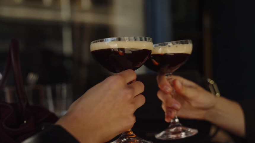 A close-up slow-motion shot of two people toasting with elegant espresso martini cocktails in a stylish bar