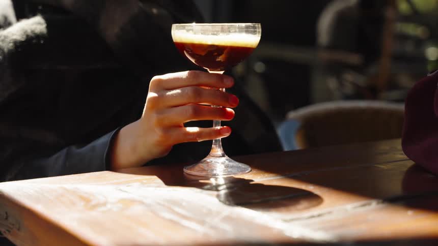 A woman lifts a glass of an espresso martini cocktail from a sunlit wooden table at an outdoor cafe