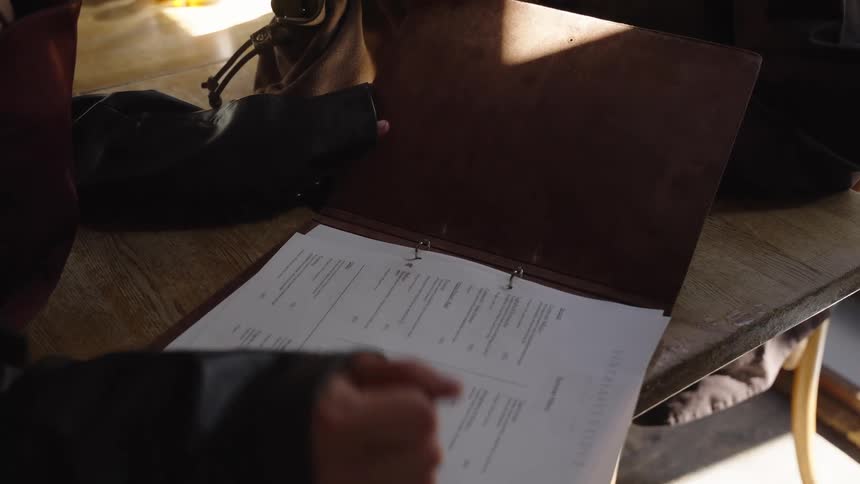 A person's hand scans and points to items on a menu inside a brown binder at a sunlit restaurant table