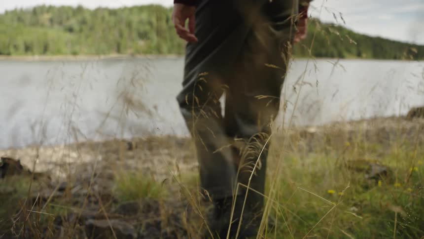 Seen through tall grass, a person wearing waders slowly walks towards the shoreline of a calm lake in the wilderness
